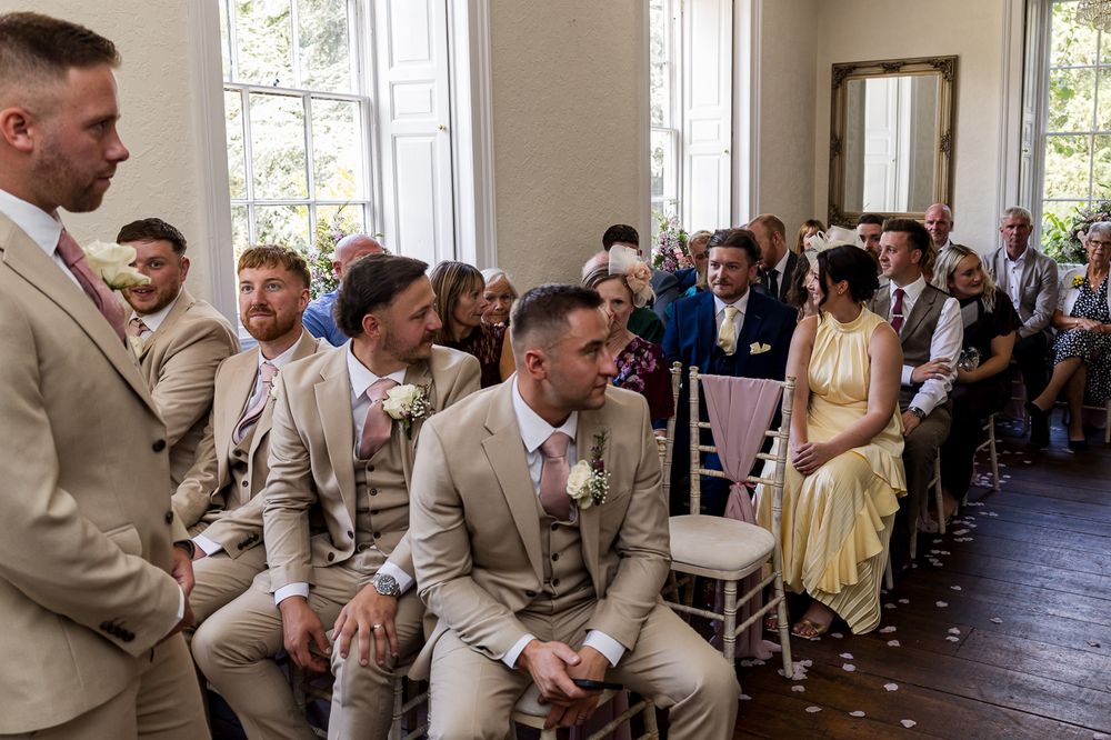 Groomsmen in tan suits seated at a wedding ceremony, with guests in the background.