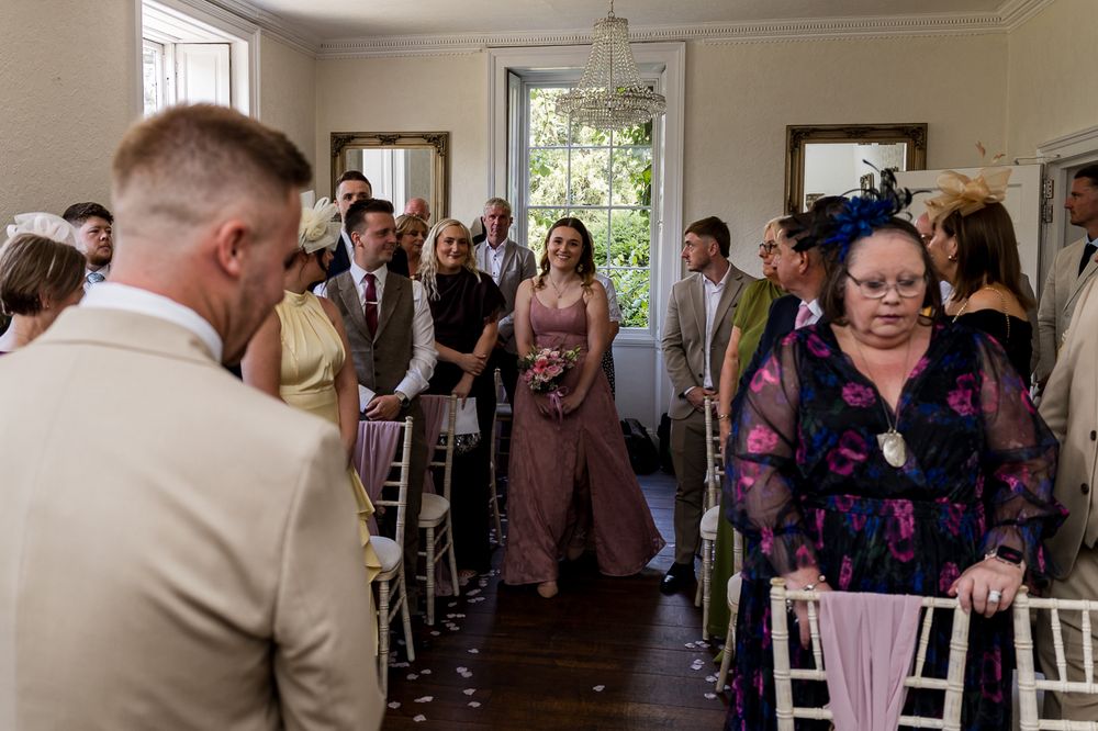 Guests walk down the aisle at an elegant indoor wedding ceremony, with a bridesmaid in pink leading the procession.