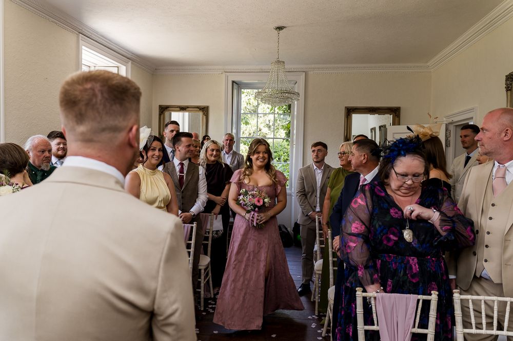 Wedding guests watch bridesmaid in mauve dress walk down aisle during indoor ceremony with white chairs.