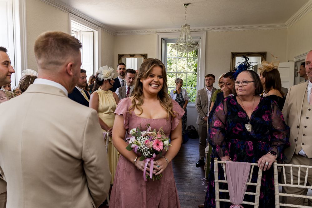 Bridesmaid in pink dress holding floral bouquet walks down aisle at elegant indoor wedding ceremony.