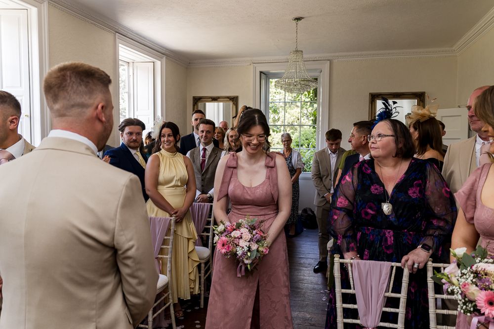 Bridesmaid in pink jumpsuit walks down the aisle holding bouquet at an elegant indoor wedding ceremony.