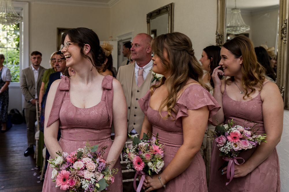 Bridesmaids in pink dresses holding floral bouquets laughing together during a wedding ceremony indoors.