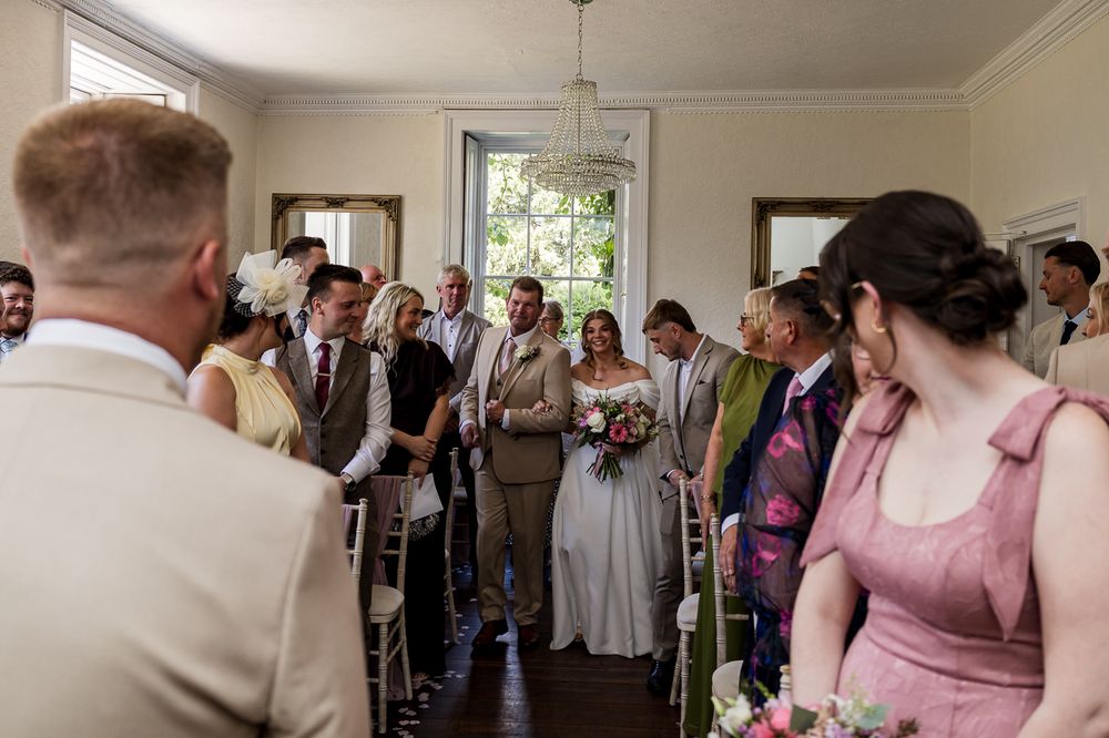 Bride walks down the aisle in a elegant gray gown, holding a bouquet, surrounded by wedding guests in a bright venue.