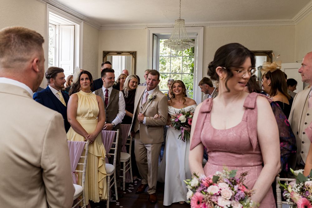 Wedding ceremony indoors with guests standing, bridesmaid in pink holding flowers, and groom waiting at the altar.