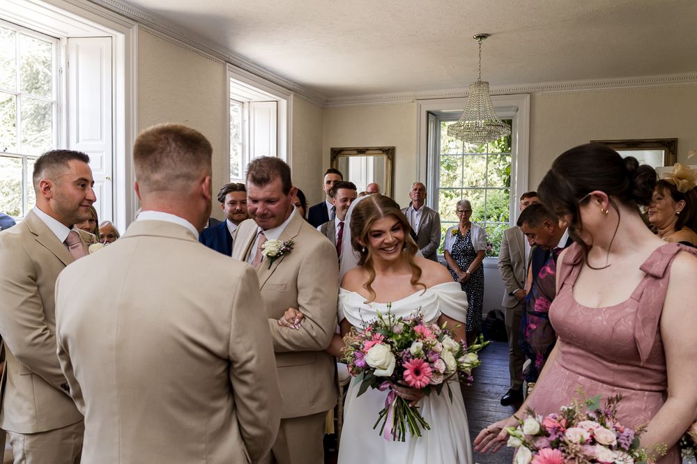 Bride in white off-shoulder gown holding pink bouquet greets guests in elegant wedding venue.