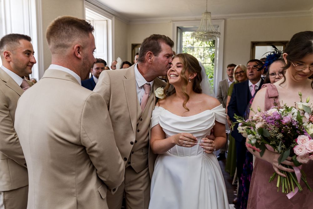 Groom kisses bride on cheek surrounded by wedding guests in elegant venue.