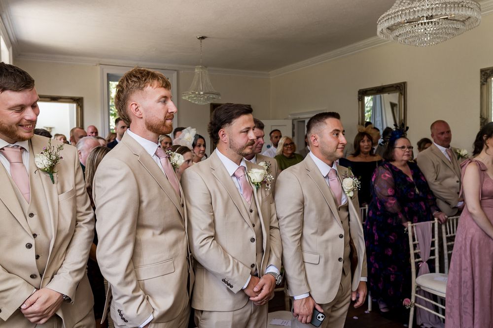 Groomsmen in tan suits with pink boutonnieres stand at wedding ceremony aisle waiting for bride.