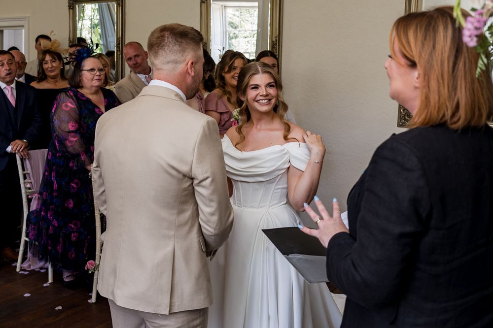 Bride smiling joyfully at groom during indoor wedding ceremony with guests and officiant in background.