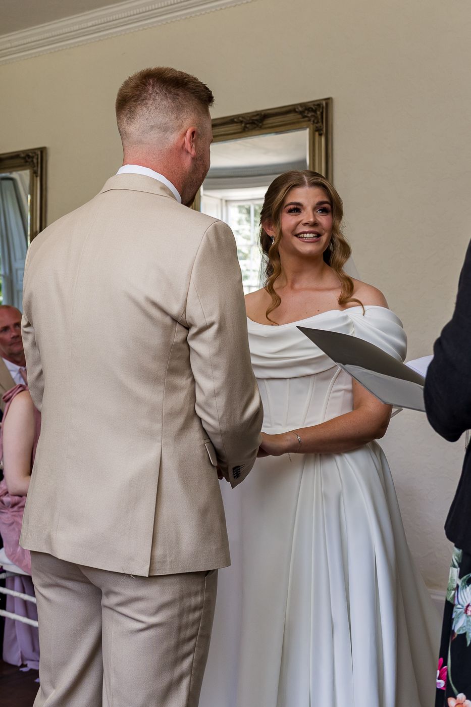Bride in off-shoulder white gown smiling at groom in beige suit during indoor wedding ceremony.