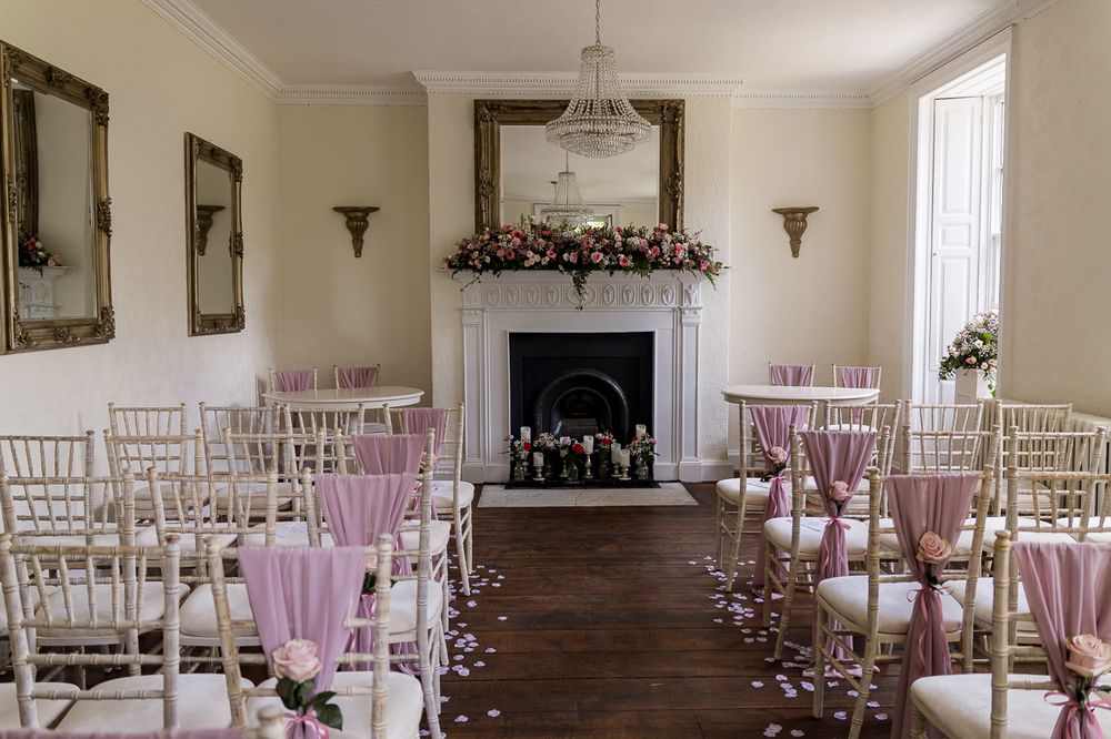 Elegant wedding ceremony room with pink chair sashes, floral fireplace display, chandelier, and rose petal aisle in a Georgian venue.