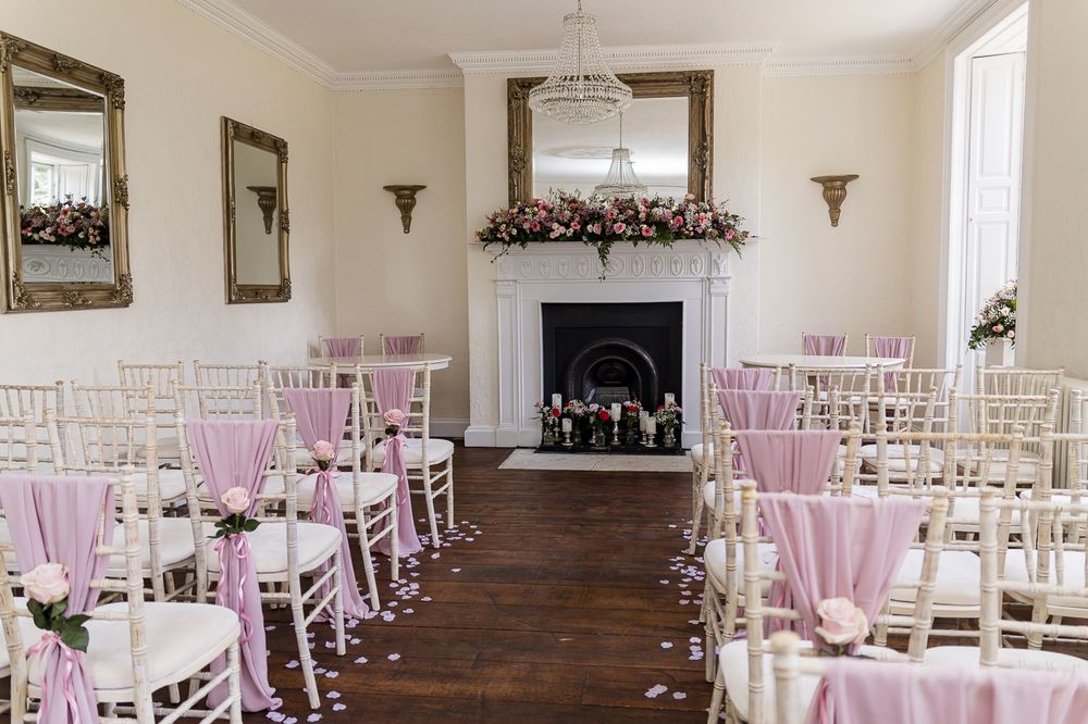 Elegant wedding ceremony room with white chiavari chairs, pink sashes, rose petal aisle, and floral fireplace mantle.