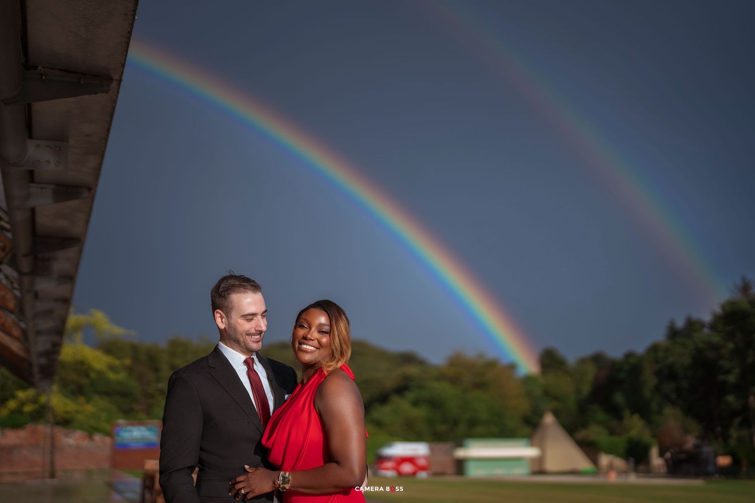 Couple embracing under a stunning double rainbow in a lush green outdoor setting, woman in red dress, man in suit.