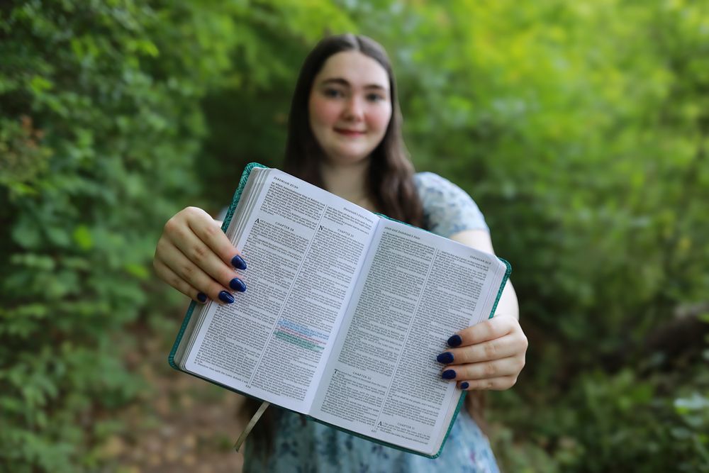Young woman holding open Bible outdoors surrounded by lush green trees, smiling with purple nail polish.