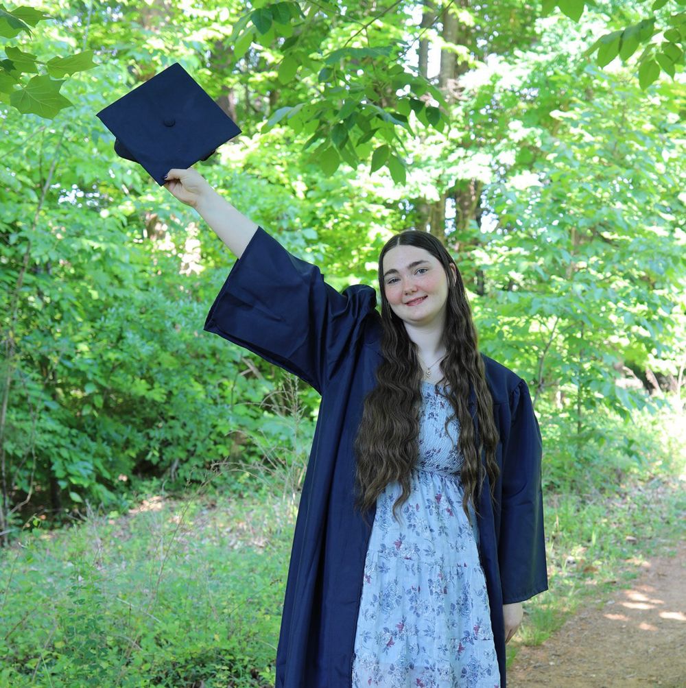 Young woman in navy graduation gown raises cap outdoors among lush green trees, celebrating academic achievement.