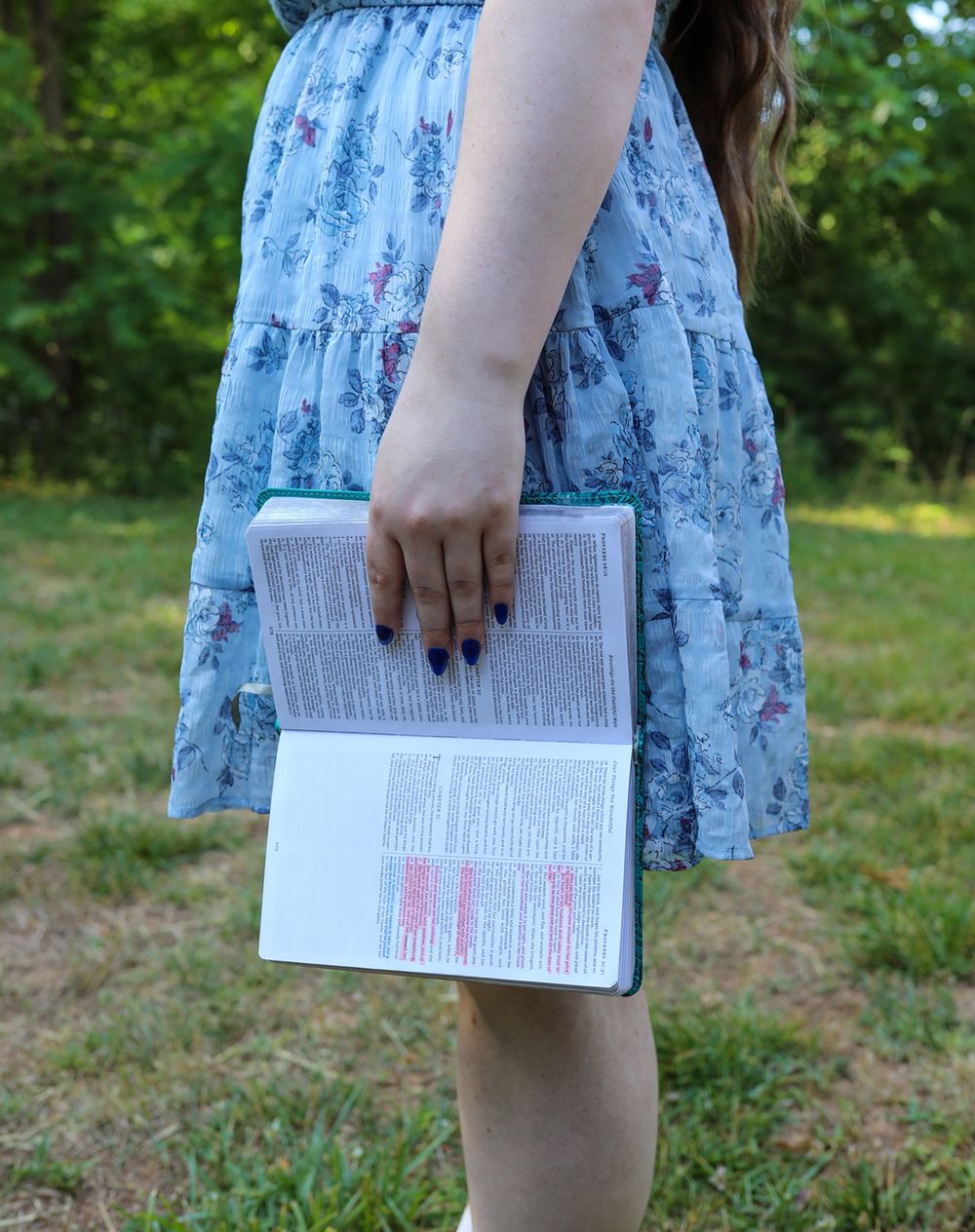 Young woman in floral blue dress holding an open Bible outdoors in a green park setting.