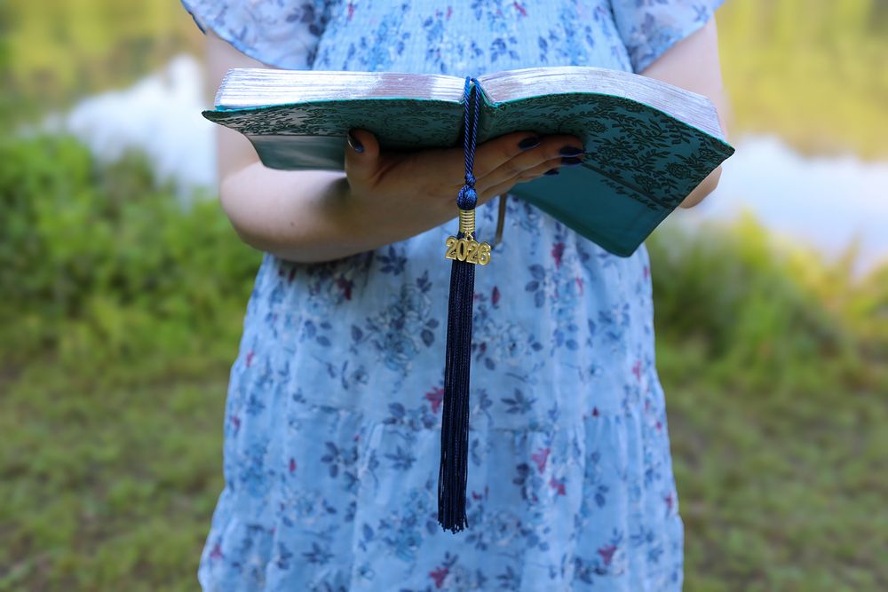 Young girl in floral dress holding an open green Quran book outdoors near water.