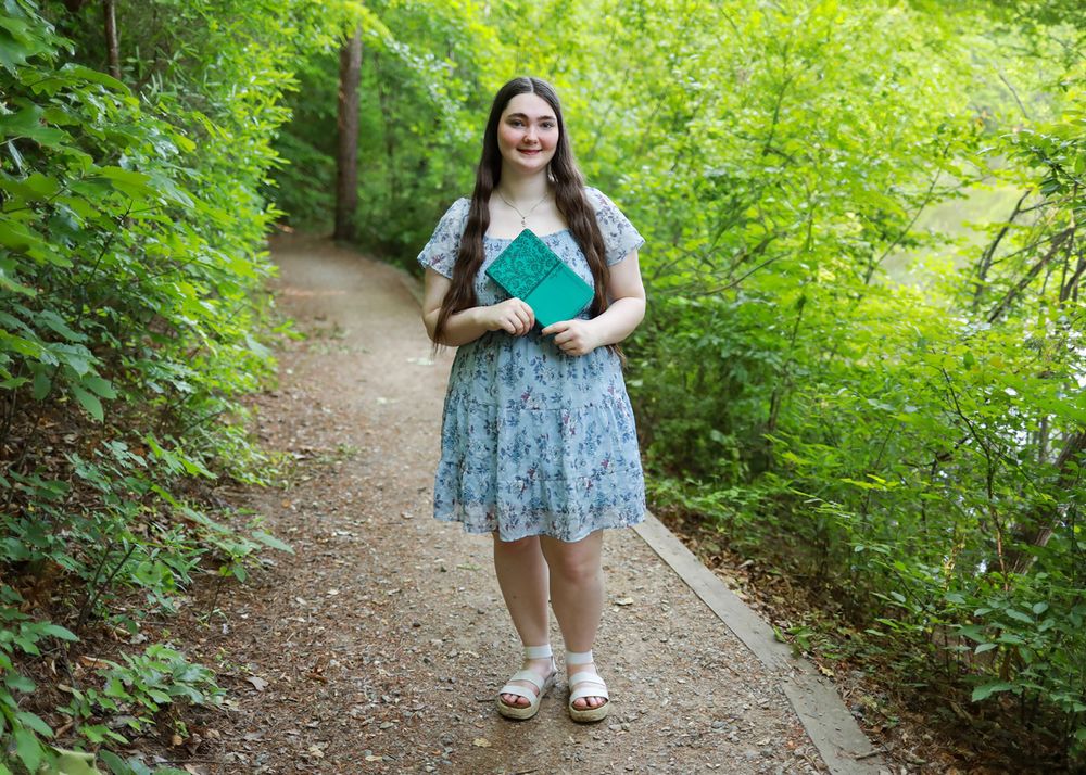 Young woman in floral dress holding green books on a scenic tree-lined garden path.