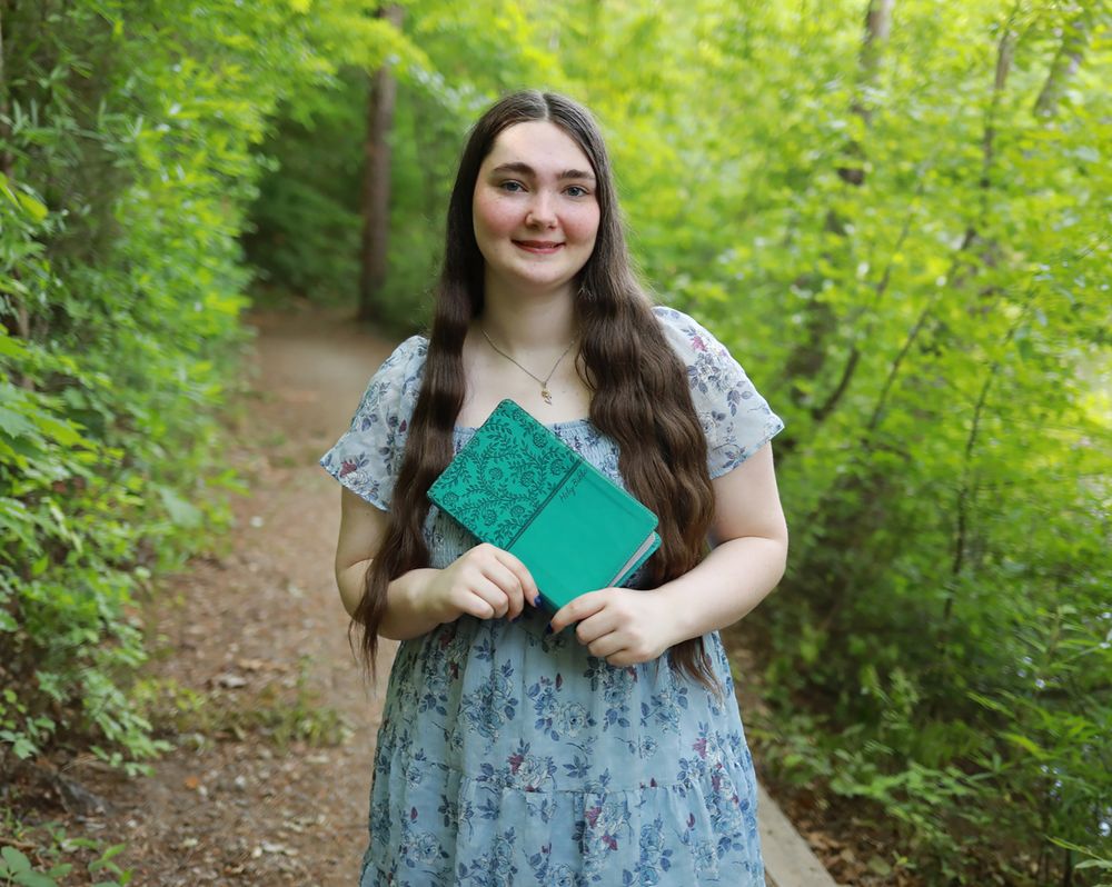 Young woman in floral dress holding green book on forest path surrounded by lush green trees.