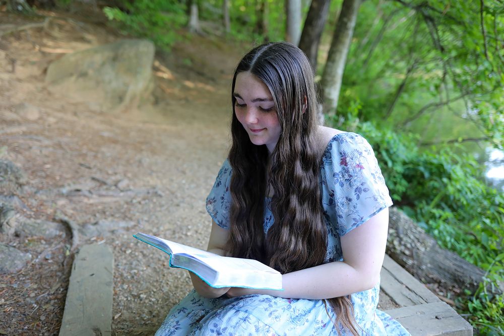 Young girl with long brown hair reading a book outdoors on stone steps surrounded by lush green trees.