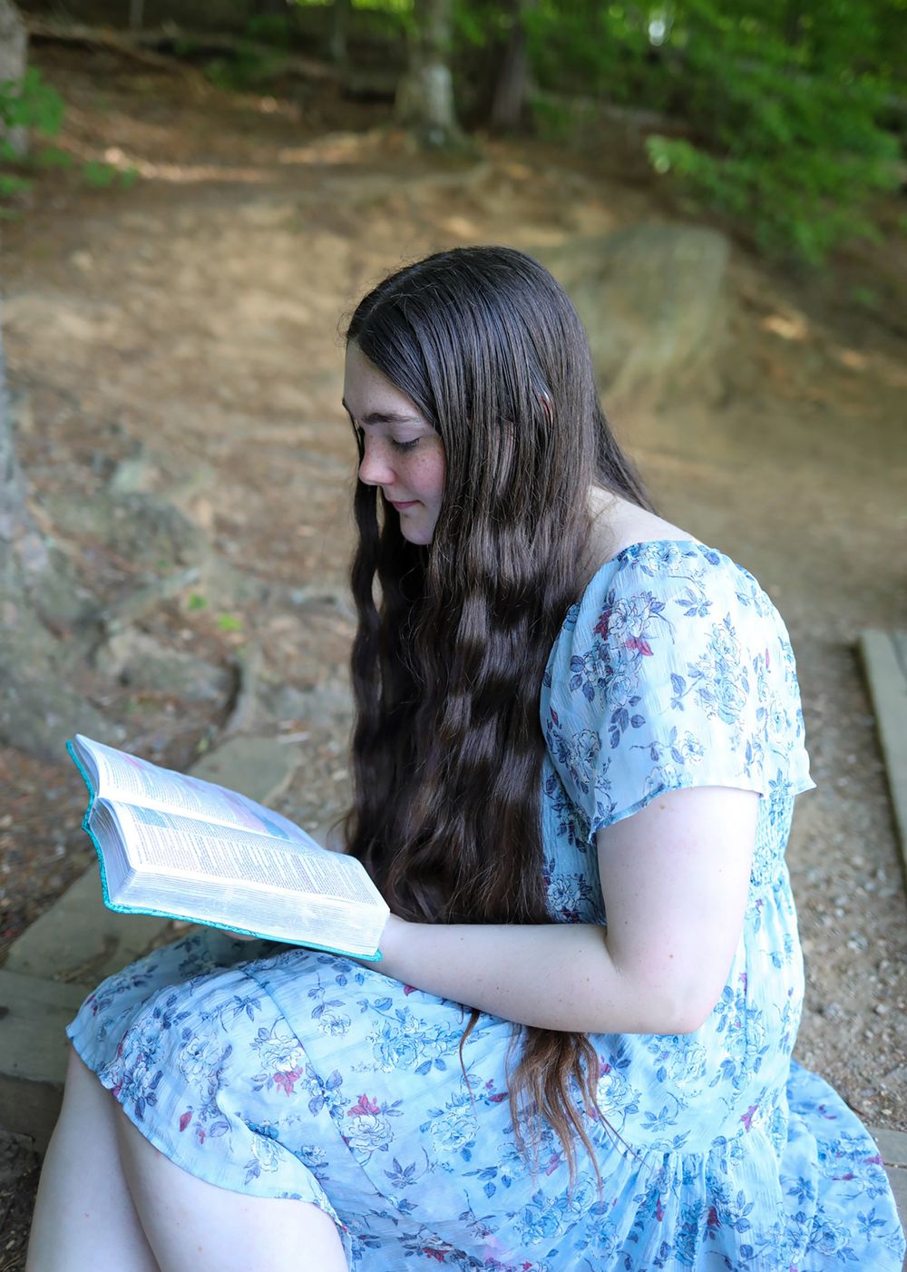 Young woman with long dark hair reads a book outdoors, wearing a floral blue dress while seated in a wooded area.