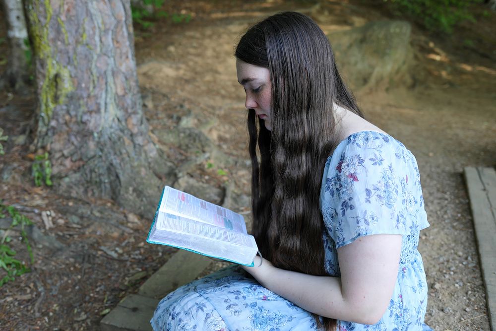 Young woman with long dark hair reading a book outdoors, seated near a tree in a floral dress.