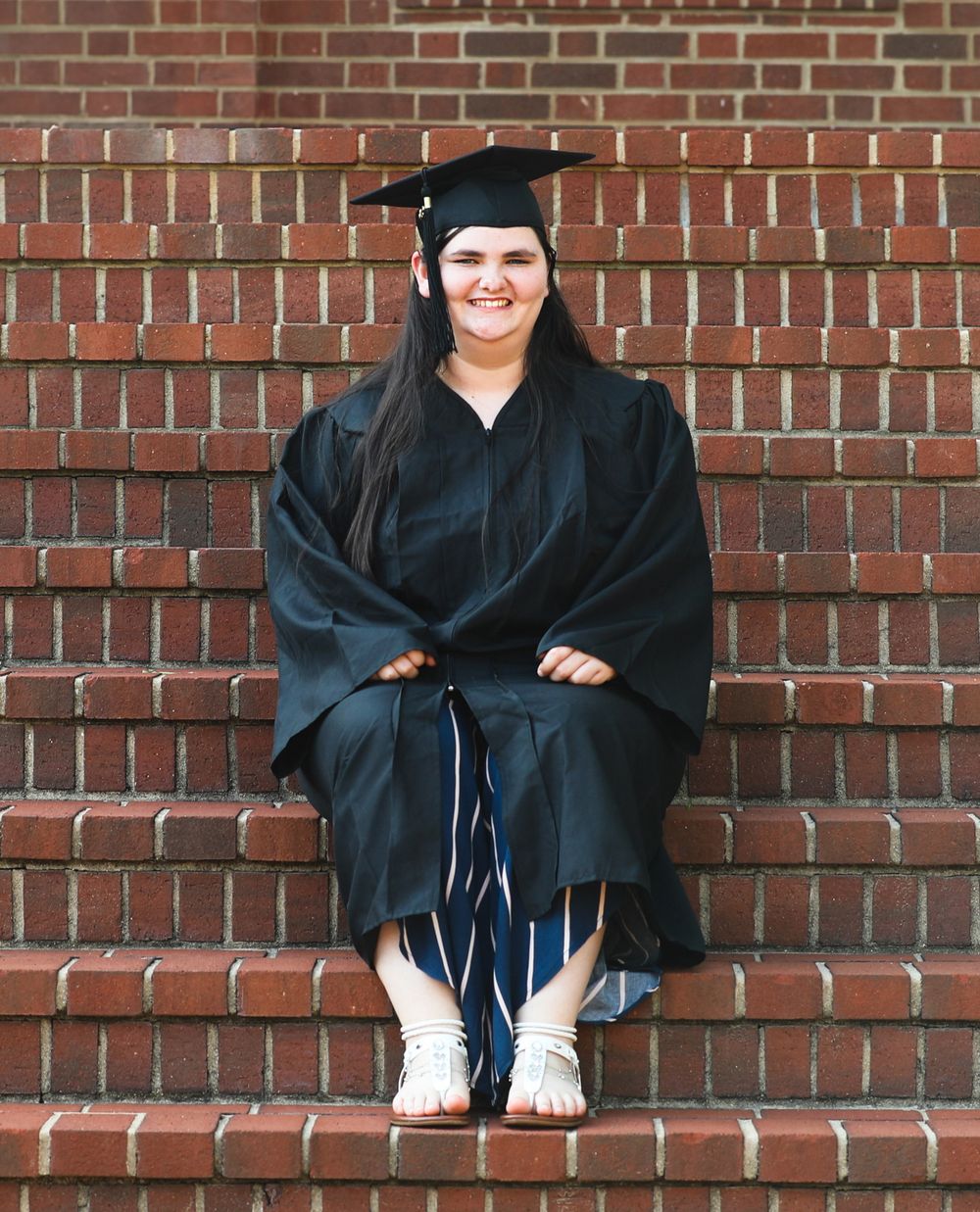 A smiling graduate in black cap and gown sits on brick steps, wearing blue striped pants and white sandals.