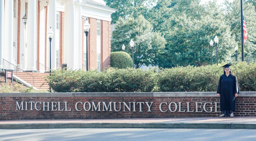 Graduate in cap and gown posing by Mitchell Community College brick sign outside campus building.