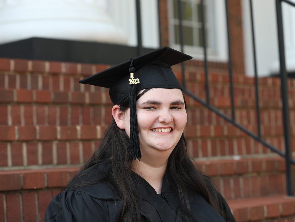 Smiling graduate in black cap and gown poses outdoors near brick steps on graduation day.