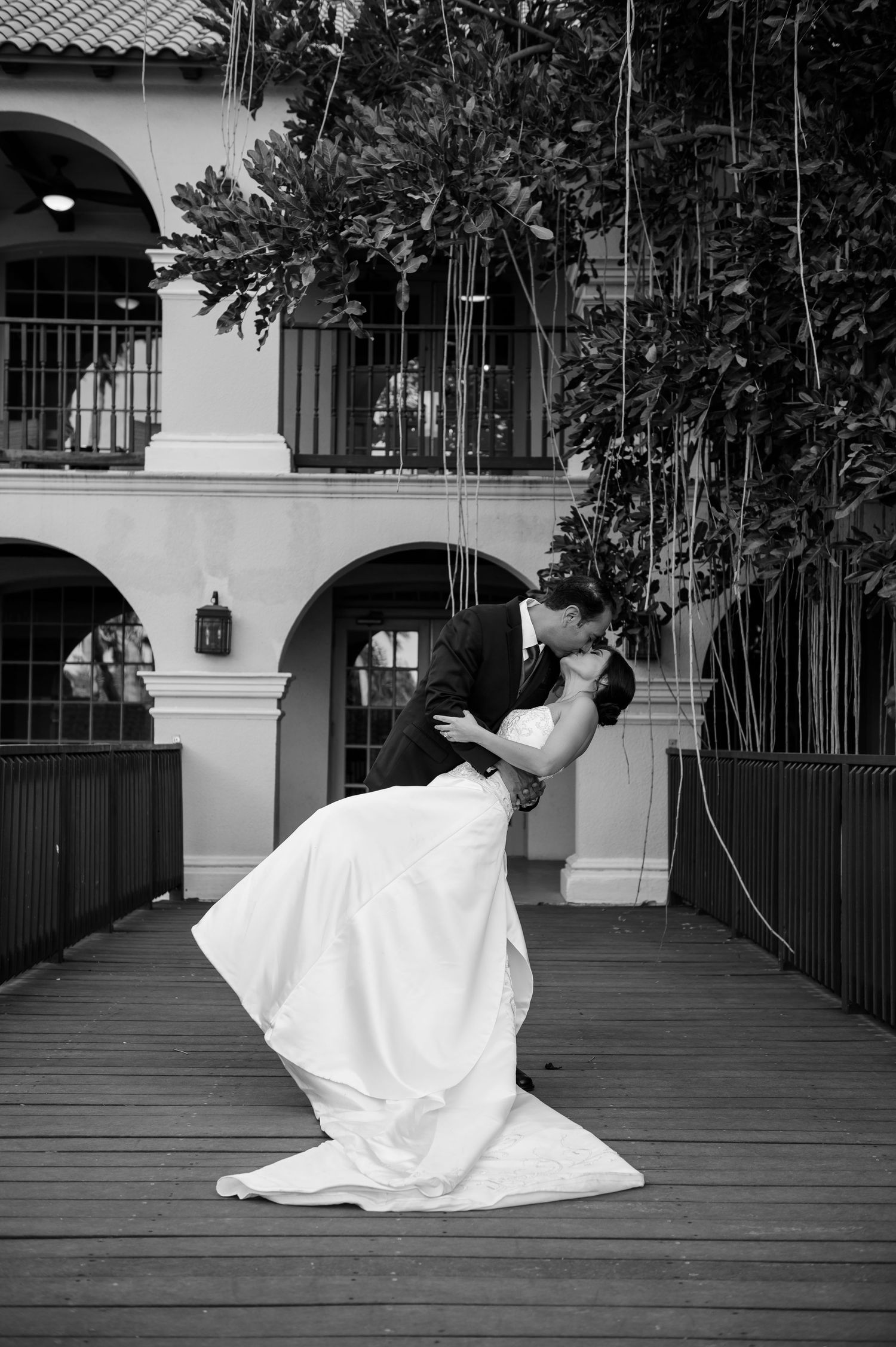 Black and white photo of a groom dipping and kissing his bride in a white gown on a wooden bridge at their wedding.