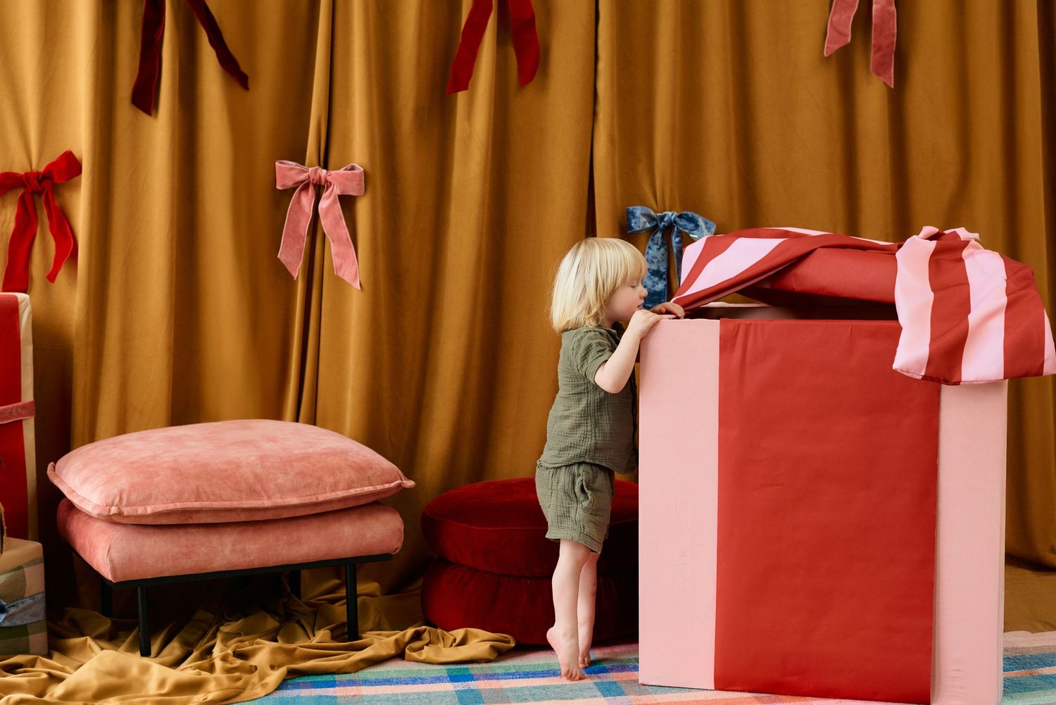 Toddler peeking inside a large red and pink Christmas gift box surrounded by festive holiday decorations.