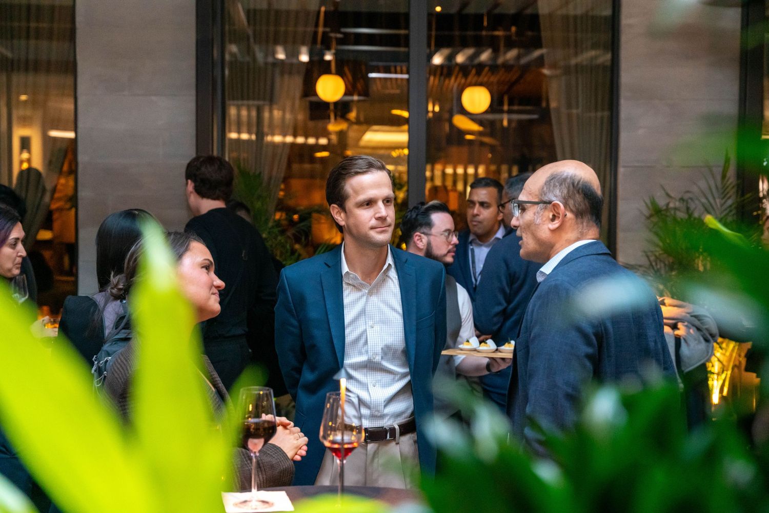 Professionals networking at an elegant indoor-outdoor cocktail reception, surrounded by lush greenery and warm ambient lighting.