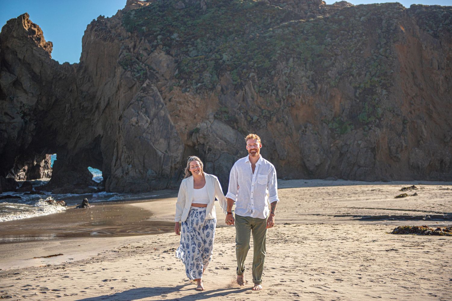 Couple holding hands walking on sandy beach with dramatic rock formations and ocean arch in background.