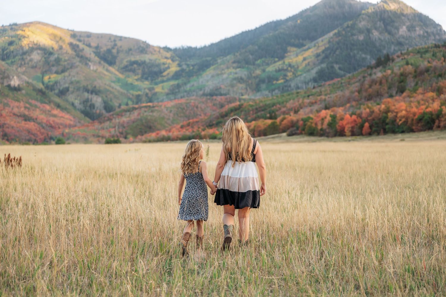 Two girls walk hand-in-hand through a golden meadow with colorful autumn mountains in the background.