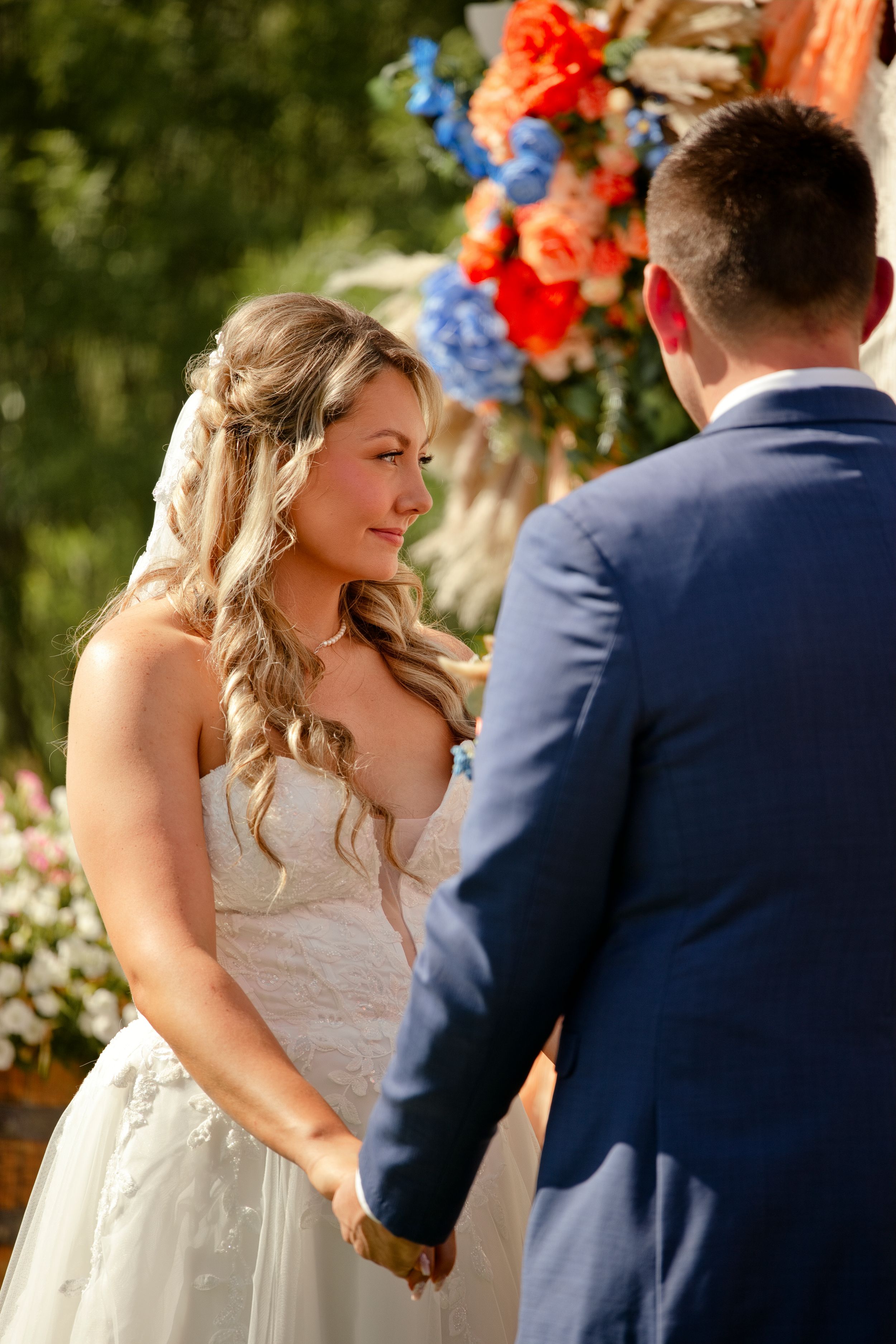 Bride smiling at groom during outdoor wedding ceremony, holding hands with colorful floral arrangement in background.