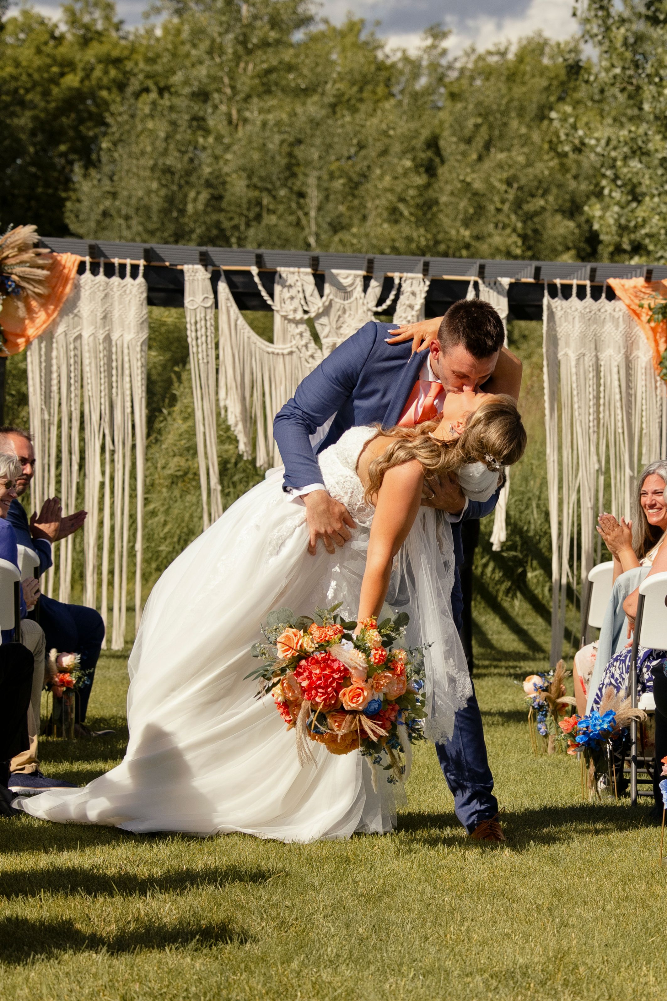 Groom dips bride for a kiss at outdoor wedding ceremony with macrame backdrop and colorful bridal bouquet.