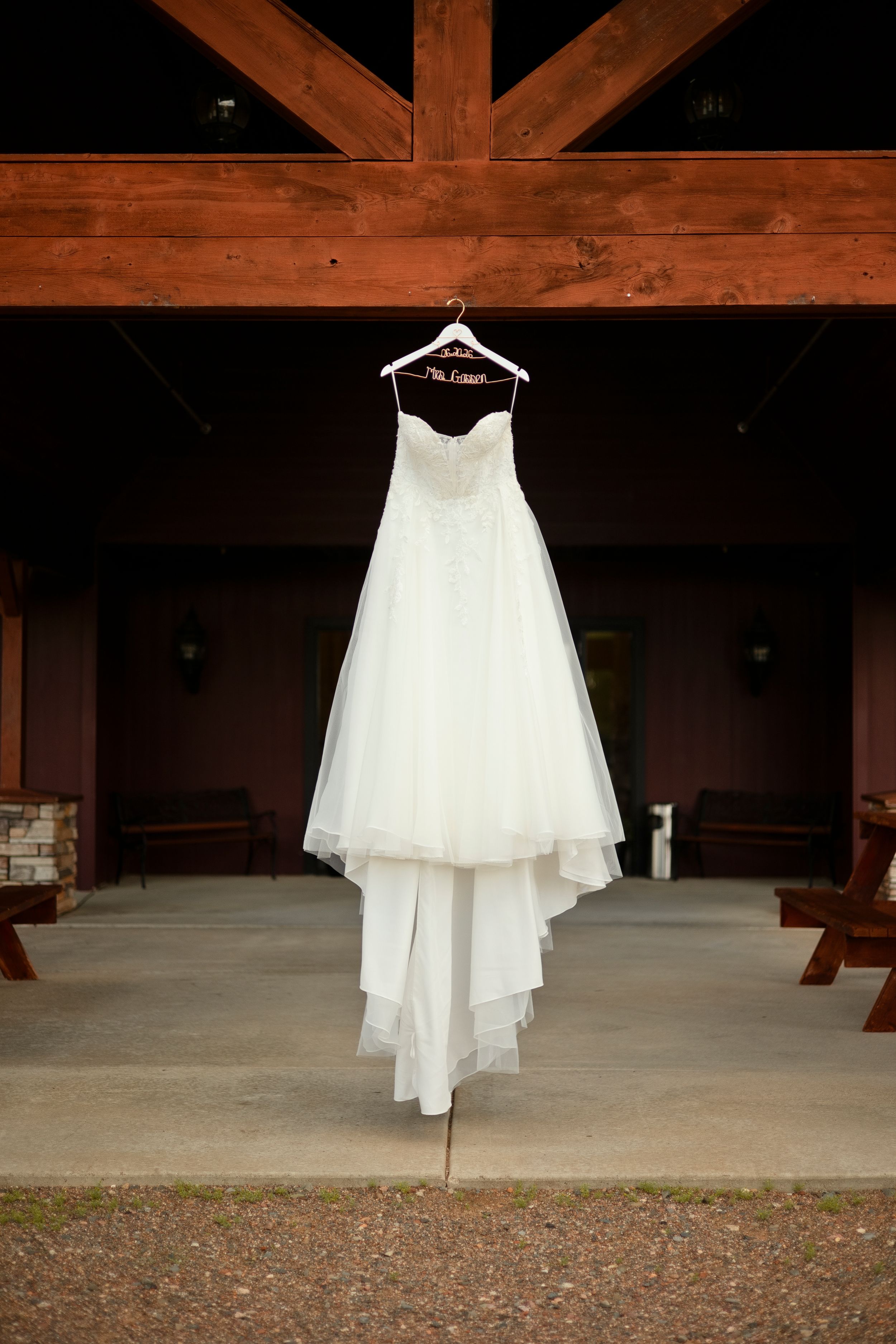 White strapless wedding dress hanging on a personalized hanger under a rustic wooden beam at an outdoor venue.