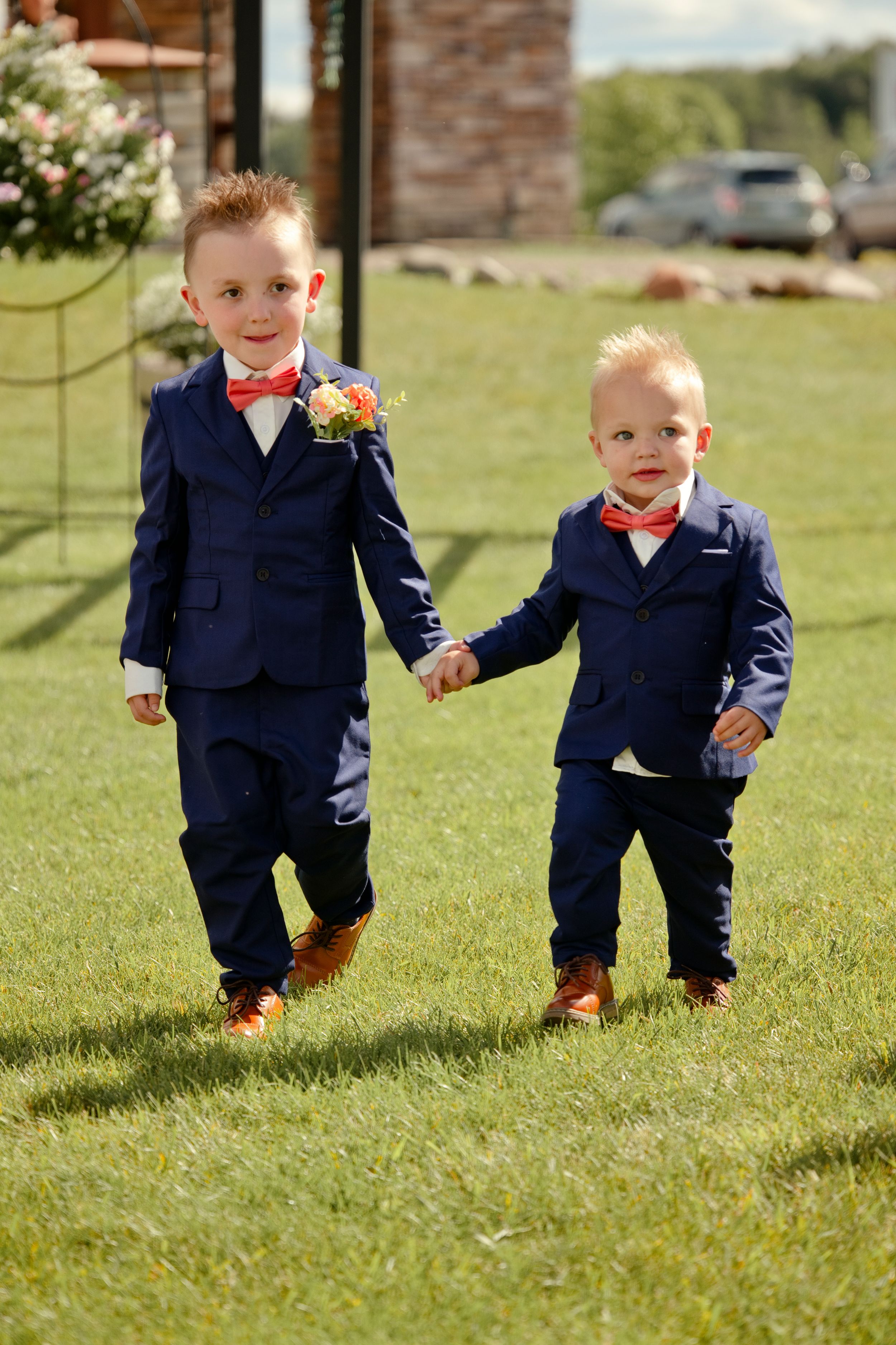 Two young boys in matching navy suits with coral bow ties and boutonnieres hold hands walking on green grass.