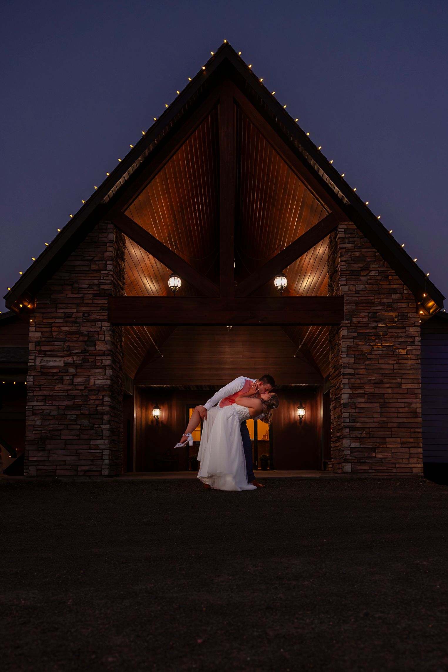Groom lifting bride in wedding attire outside a stone chapel with string lights at dusk.