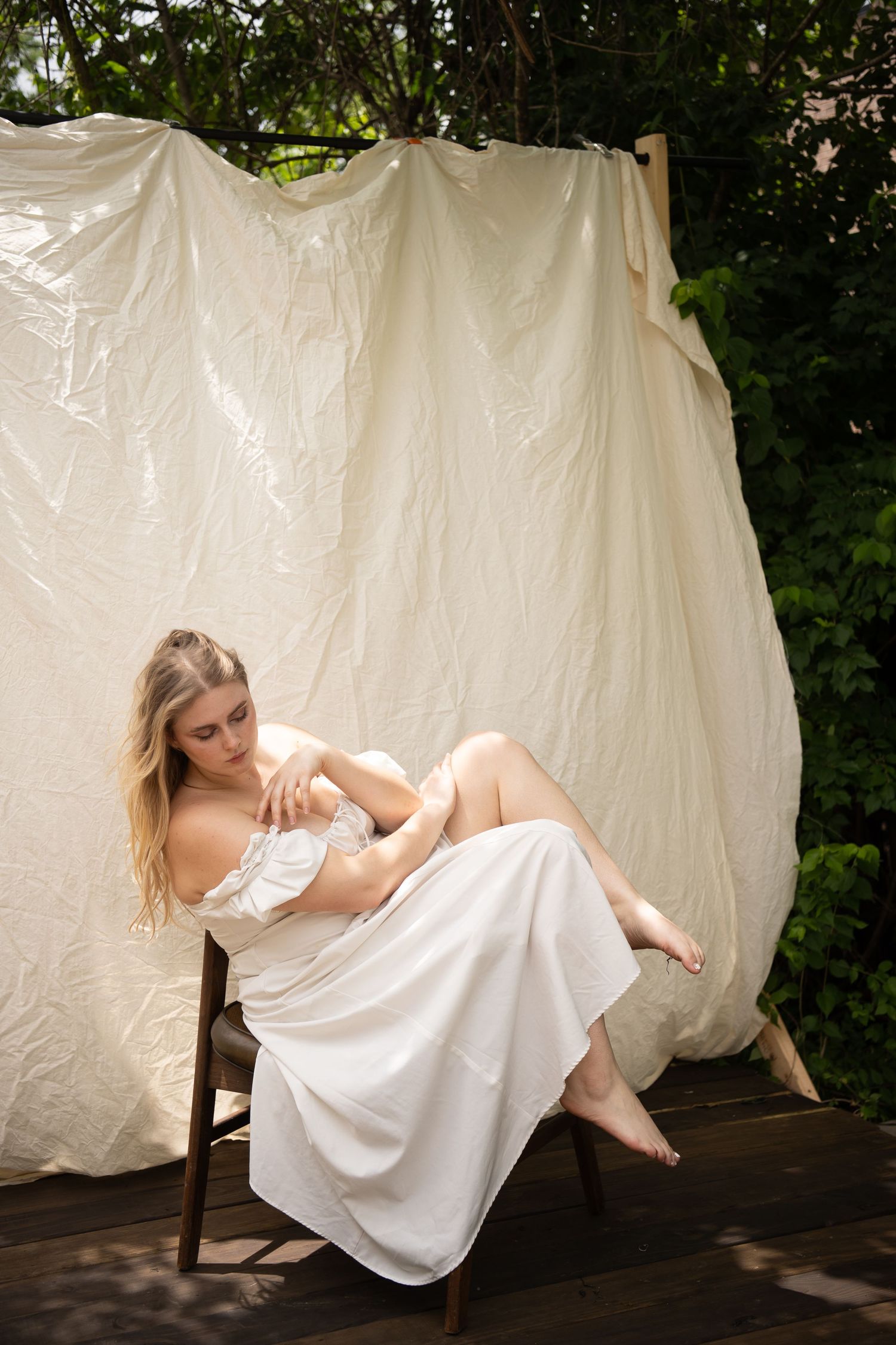 A blonde woman in a white off-shoulder dress sits barefoot on a wooden chair against a draped fabric backdrop outdoors.