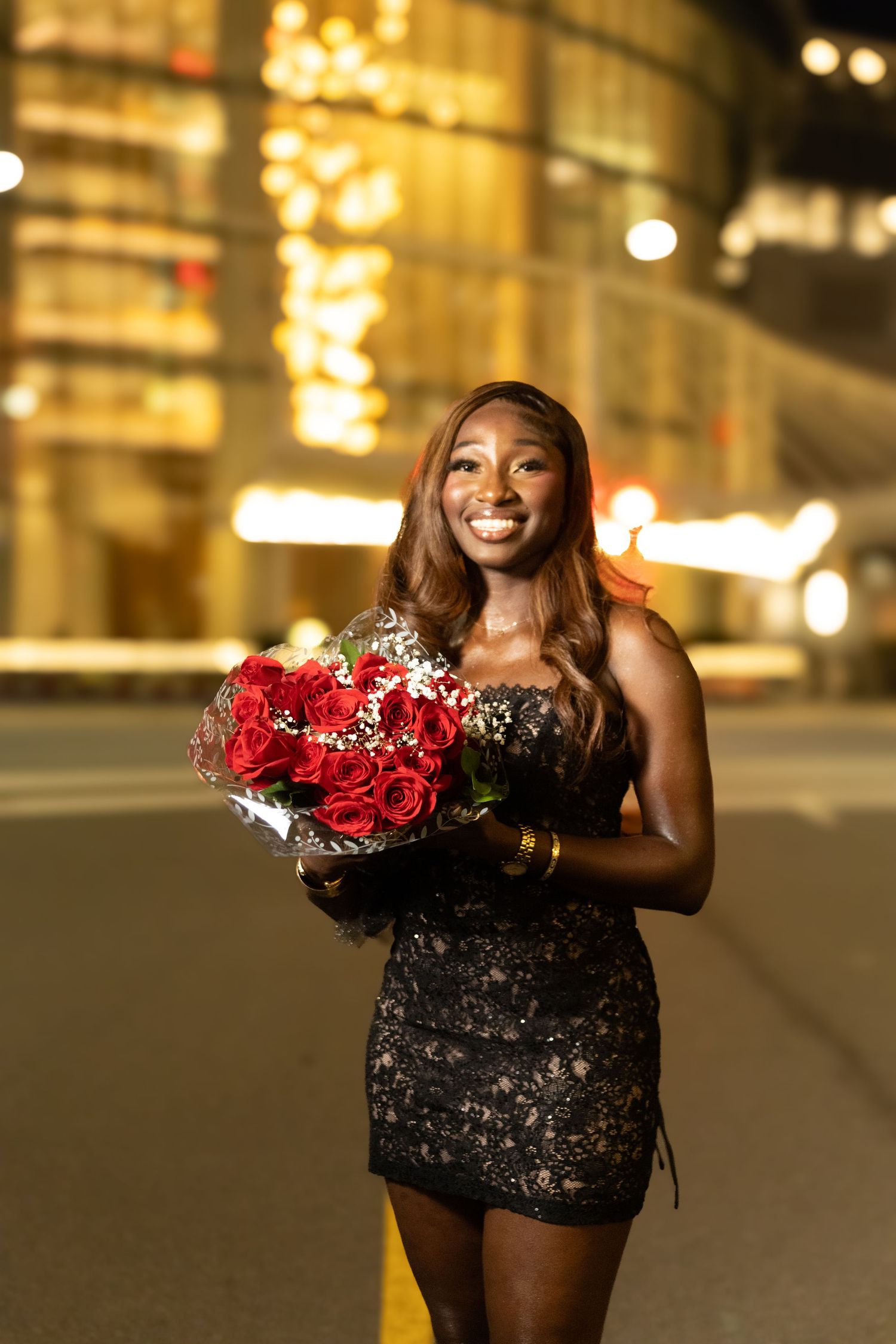 Smiling woman in black dress holding red roses bouquet outdoors at night with city lights in background.
