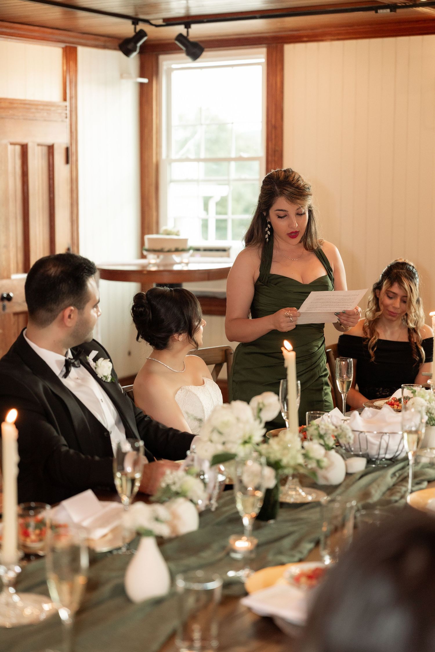 Groom in black tuxedo raises champagne glass for wedding toast with guests at elegantly decorated reception dinner table.