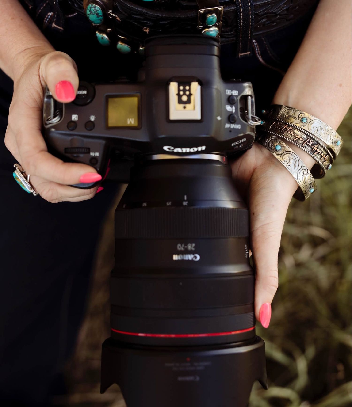 Close-up of hands with pink nails and silver turquoise bracelets holding a Canon mirrorless camera with a large lens.