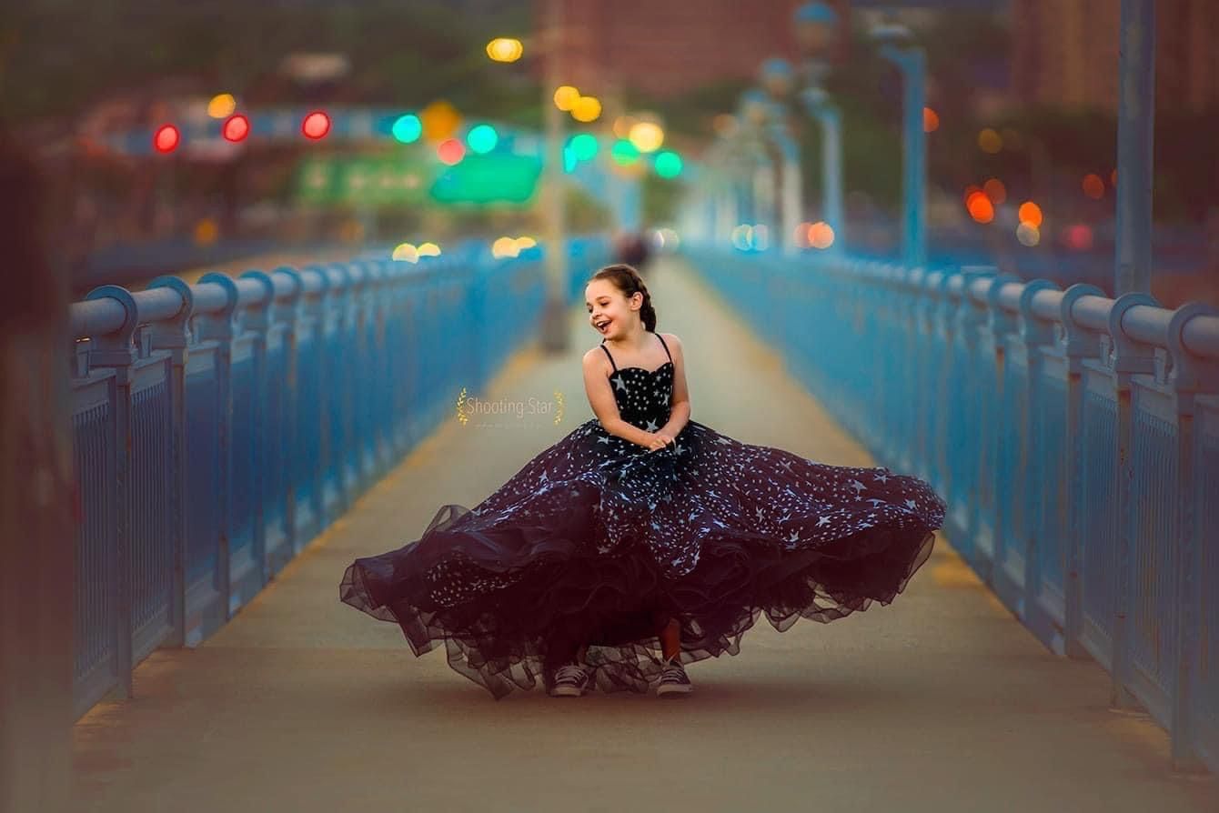 Smiling girl in a sparkling black tulle dress twirls on a blue-railed pedestrian bridge at dusk with colorful bokeh city lights behind her.