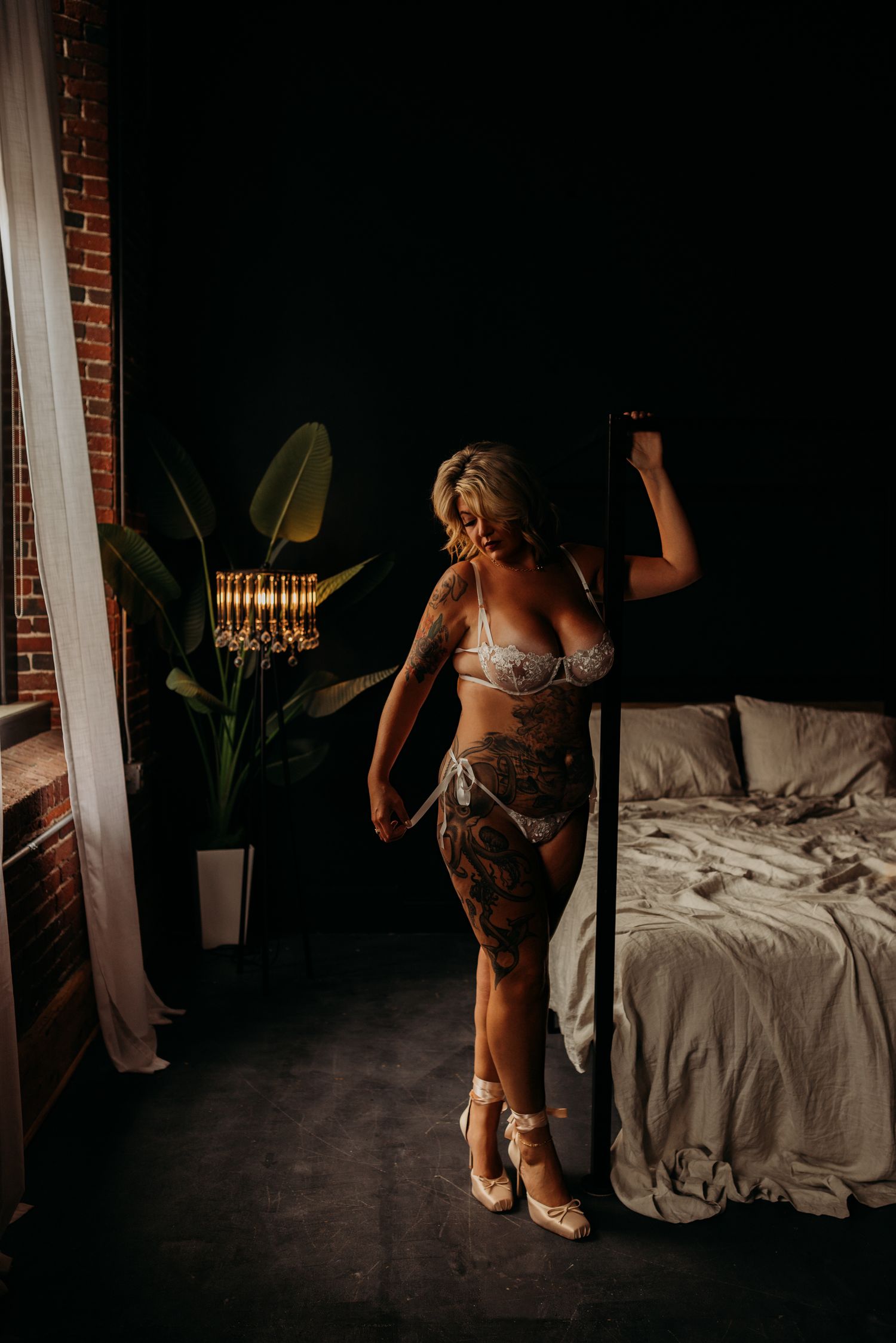 Blonde woman in lace lingerie and heels poses beside a bed in a moody, dimly lit boudoir studio.