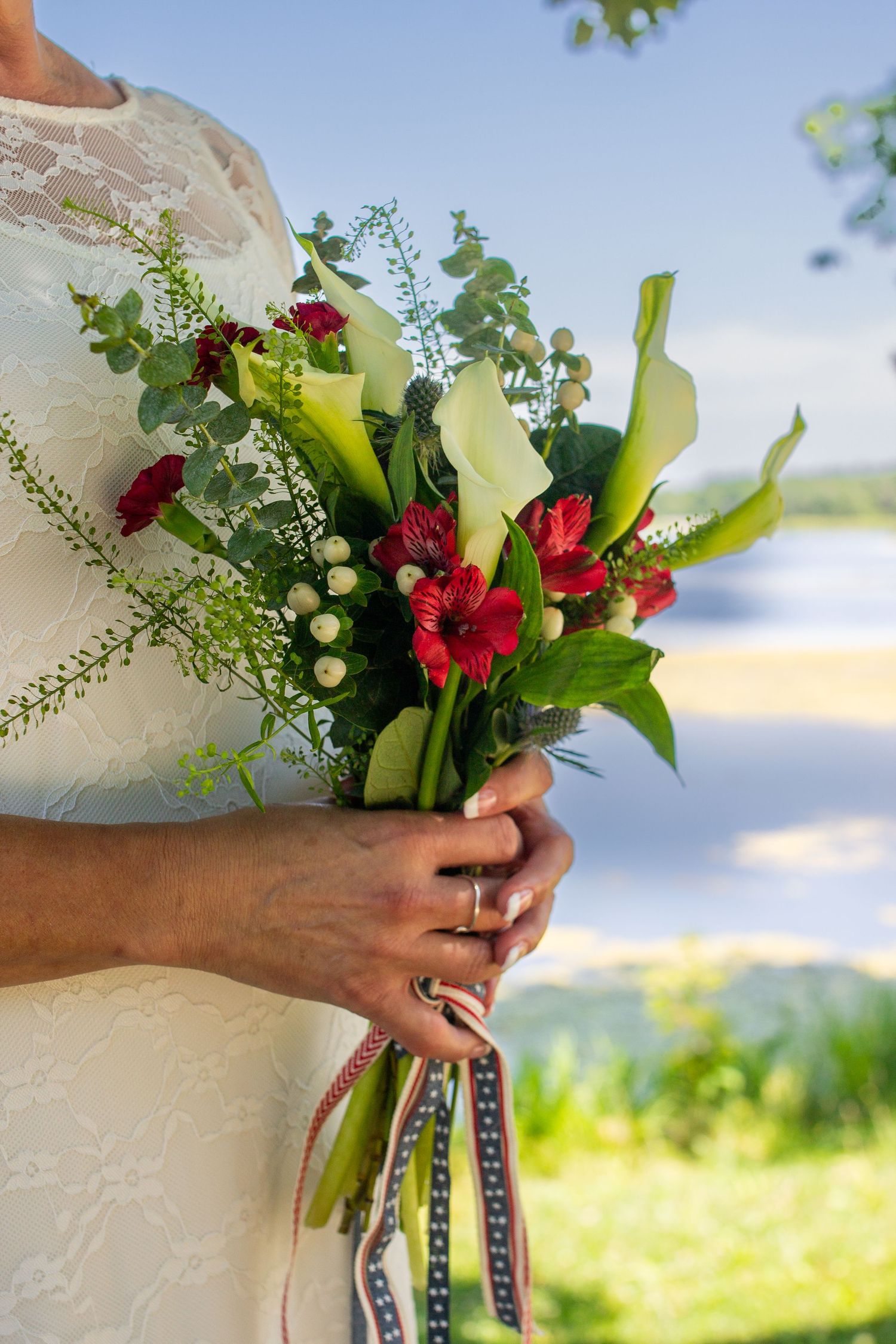 Bride holding a colorful wedding bouquet with red gerberas, calla lilies, and greenery near a serene lakeside backdrop.
