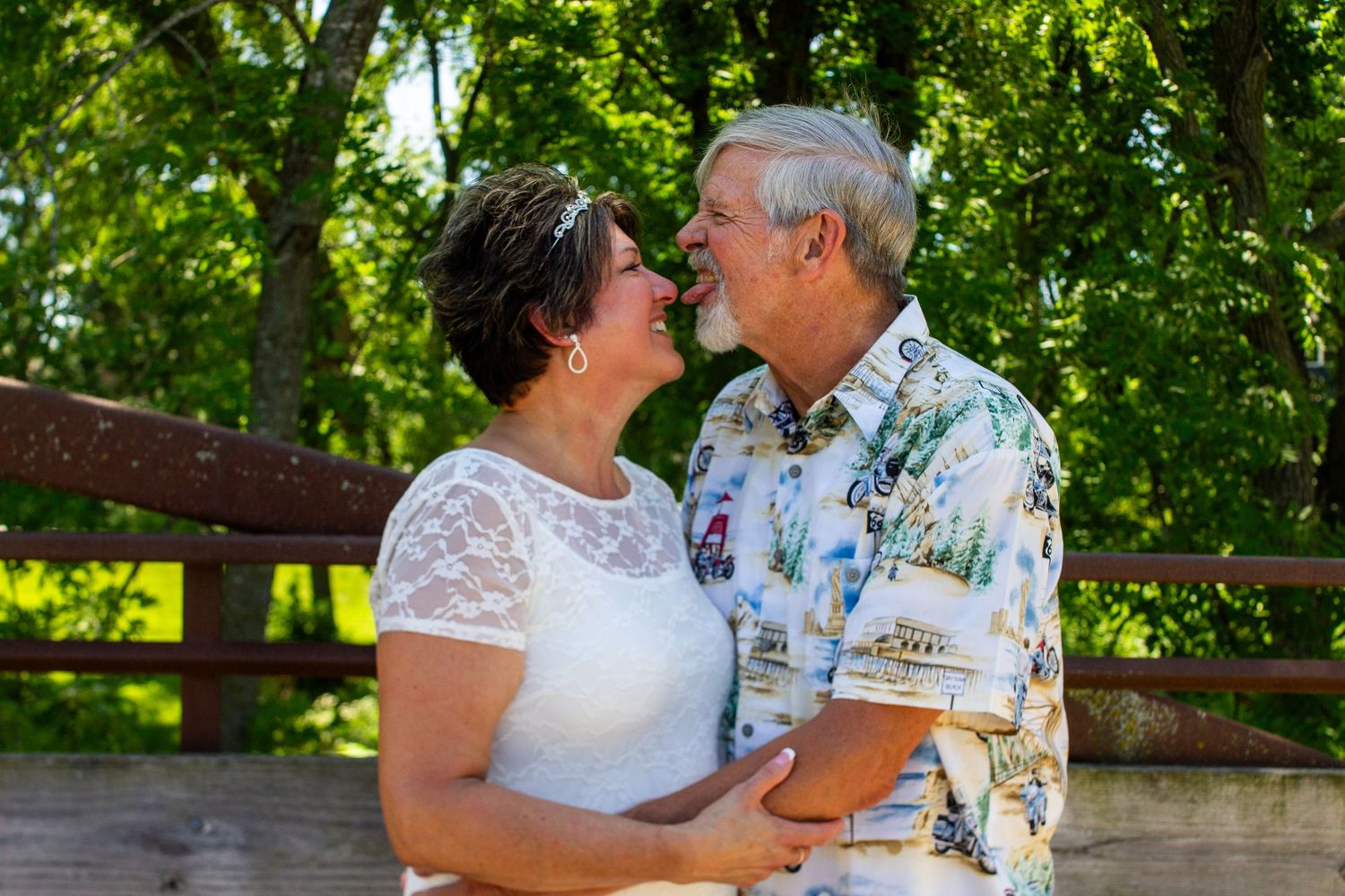 Older couple sharing a romantic moment outdoors near a wooden bridge, woman in white lace dress, man in floral shirt.
