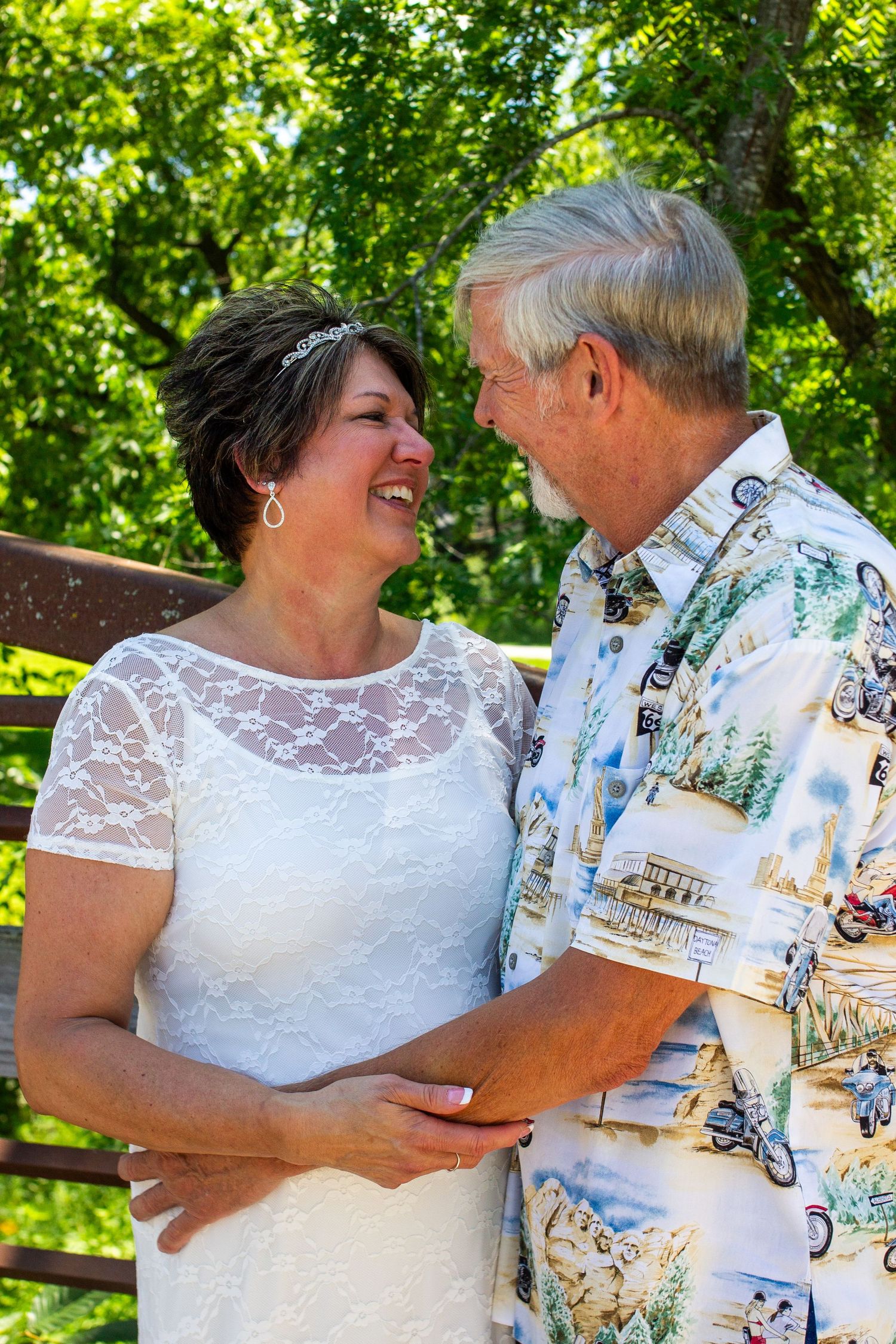 A smiling couple in wedding attire embraces outdoors, the bride in white lace and groom in a Hawaiian shirt, surrounded by green trees.