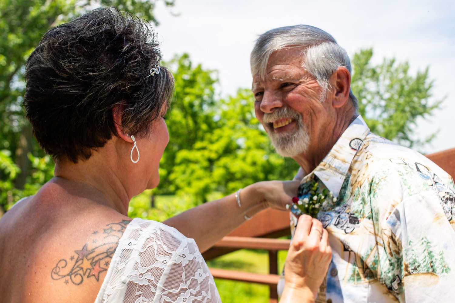 Older couple smiling outdoors, woman adjusting man's floral bow tie at a wedding celebration.