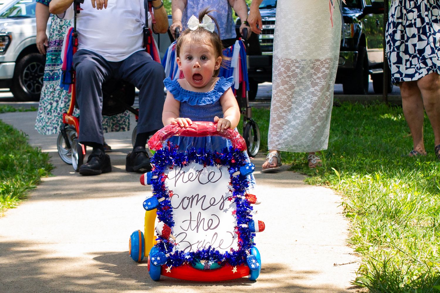 Toddler girl pushes patriotic red, white, and blue decorated walker with 'Here Comes the' sign in outdoor parade.