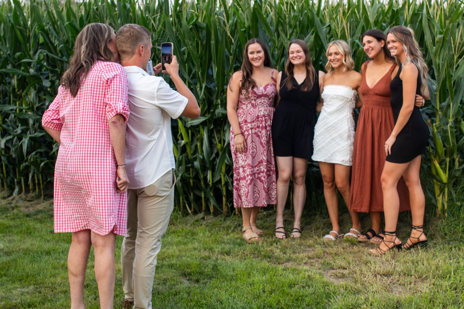 A photographer captures a group of five young women posing in front of a lush green cornfield on a sunny day.
